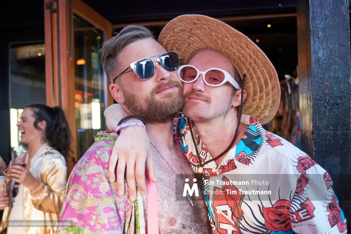 Two friends share an intimate moment at White Owl Social Club during Your Sunday Best's season opener in Portland, Oregon. The bearded man in reflective sunglasses embraces his companion, who sports white oval sunglasses and a woven straw hat alongside a vibrant floral Hawaiian shirt. Golden afternoon light filters through the venue's windows, creating a warm, celebratory atmosphere as other partygoers mingle in the background of this popular summer day party.