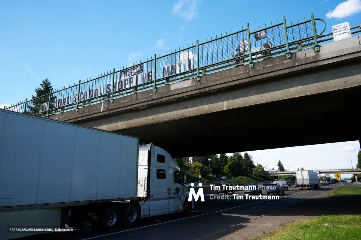 Activists from the Indivisible movement have suspended a protest banner reading "No Work School Shopping May 1st" across a concrete overpass spanning Interstate 5 in North Portland's Humboldt neighborhood. Below, a white semi-truck and steady stream of traffic flow beneath the weathered bridge structure, while afternoon sunlight filters through wispy clouds above. The grassroots demonstration transforms mundane infrastructure into a platform for labor organizing, capturing the intersection of highway commerce and social activism.