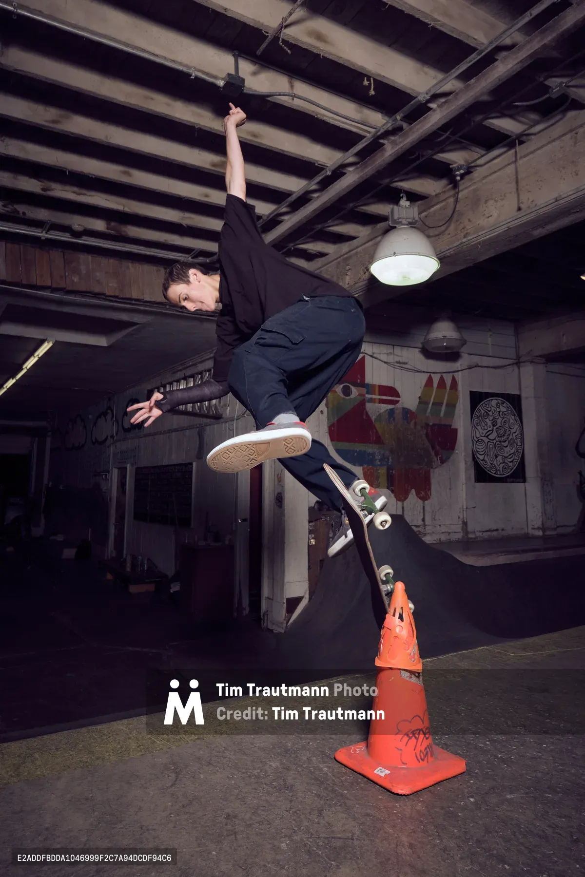 A skateboarder launches into a gravity-defying kickflip above a graffitied orange traffic cone in the industrial confines of Commonwealth Skateboarding in Portland's Buckman neighborhood. The raw concrete floor and exposed ceiling beams create a gritty backdrop, while weathered murals and pendant lighting cast dramatic shadows across this underground skating sanctuary. The athlete's black attire contrasts sharply against the weathered wooden walls, capturing a moment of pure athletic poetry in motion.