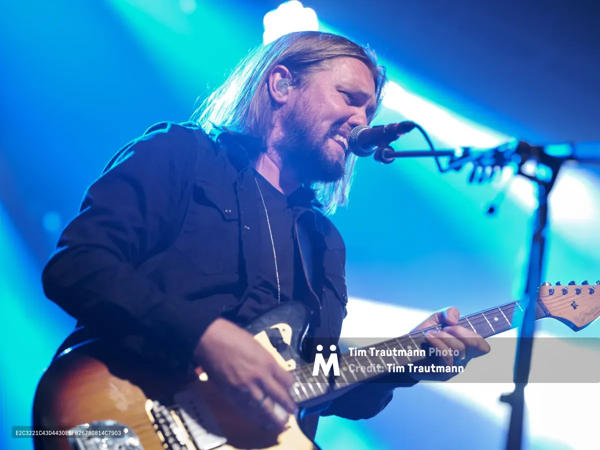 Russell Marsden, vocalist and guitarist of Band of Skulls, delivers an intense performance during the band's 10 Year Anniversary Tour at Webster Hall in Manhattan. Bathed in dramatic blue and turquoise stage lighting, Marsden leans into his microphone with passionate intensity while playing his electric guitar. His shoulder-length hair catches the ethereal glow as beams of light pierce through the atmospheric haze, creating a cinematic moment of rock performance art.