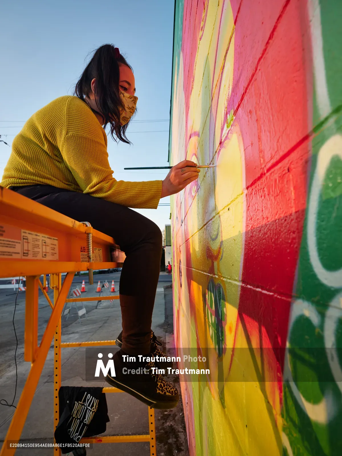 Multidisciplinary artist Vilatose works methodically on her mural 'Jardín Cubana' depicting three female figures, perched on an orange scissor lift against the expansive wall of a building in Portland's Alberta Arts District. Golden hour light bathes the scene as she adds delicate details to the rainbow-hued artwork featuring coral and yellow paint strokes, her leopard print face mask and mustard sweater creating a warm counterpoint to the vivid greens and pinks cascading across the concrete canvas. The industrial lift and scattered traffic cones below speak to the organized chaos of public art creation in this culturally rich Northeast Portland neighborhood.