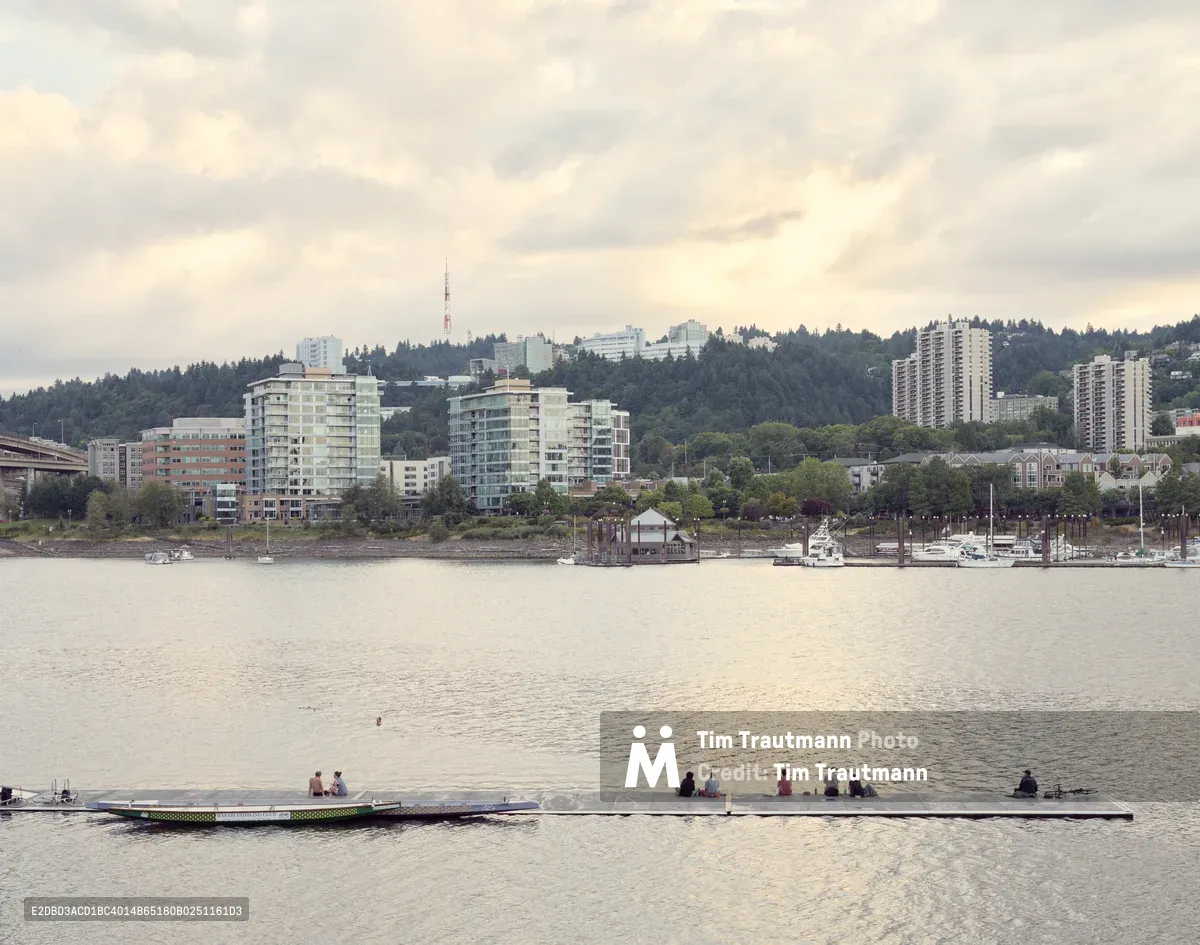 People enjoy the early evening at Holman Dock as soft light reflects across the Willamette River's surface. The forested hills of Southwest Portland rise in the background, crowned by the distinctive towers of Oregon Health and Science University and scattered residential high-rises. Moored sailboats dot the marina while dramatic clouds gather overhead, creating a quintessential Pacific Northwest waterfront scene.
