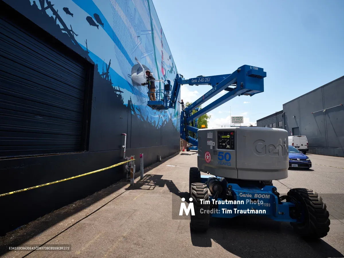 Jeremy Nichols and Devin Finley collaborate on a large-scale street art installation in Southeast Portland's Central Eastside district, suspended in a blue Genie boom lift against the industrial backdrop. The artists work methodically on their vibrant mural featuring flowing blues and organic forms, transforming the stark warehouse wall under the clear afternoon sky. Yellow caution tape cordons the work area while the mechanical lift provides steady access to the upper reaches of their canvas.