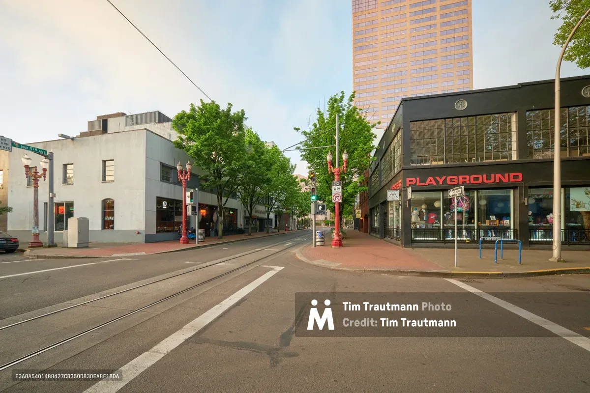 Empty asphalt stretches through Portland's Old Town Chinatown district on Northwest Couch Street, where red ornamental lamp posts stand sentinel along the pedestrian median. The PLAYGROUND storefront anchors the right side of the frame, its glass block facade catching the soft evening light, while a golden high-rise tower looms in the distance beyond verdant street trees. This tranquil urban scene captures the neighborhood's blend of cultural heritage and modern development.