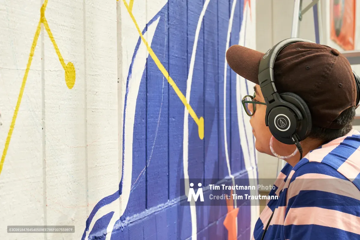 Mexican-American artist Maria Rodriguez, known as Sparkykneecap, focuses intently on her vibrant mural "Let's Talk" at Open Signal in Portland's Eliot neighborhood. Wearing Audio-Technica headphones and a navy striped shirt, she works on bold purple and yellow geometric forms painted on concrete walls. The commissioned piece explores themes of identity and culture while maintaining her signature playful aesthetic, bringing color and conversation to this Northeast Martin Luther King Jr. Boulevard arts venue.