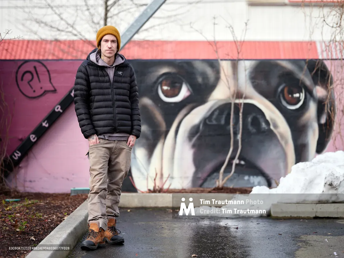 Artist Wetiko stands confidently before his striking mural of a pug's face at the Clay Creative Building in Southeast Portland's Hosford-Abernethy neighborhood. The dramatic close-up of the dog's expressive brown eyes and wrinkled snout dominates the pink wall behind him, creating a powerful juxtaposition with the artist's contemplative pose. Winter light filters through bare branches overhead, casting a subdued atmosphere over the scene while patches of snow dot the urban landscape.