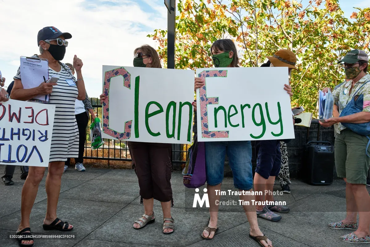 Masked protesters gather on a sun-dappled sidewalk along McLoughlin Promenade in Oregon City, holding handmade signs advocating for clean energy outside Representative Kurt Schrader's office. The demonstrators, dressed in casual autumn attire, stand beneath trees showing the first hints of fall color against a partly cloudy sky. Their collective presence creates a peaceful yet determined tableau of grassroots environmental activism in this small Oregon community.