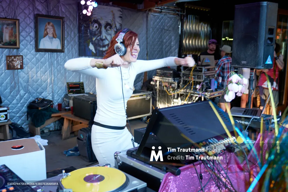 DJ Doctor Dakar spreads her arms wide in triumphant celebration behind the turntables at White Owl Social Club during the Your Sunday Best season opener in Portland. The red-haired performer, adorned with professional headphones and a crisp white top, commands attention against the venue's quilted silver walls decorated with eclectic portrait photography. Purple and yellow lighting bathes the DJ booth while vinyl records and mixing equipment create a vibrant sonic landscape around her energetic performance.