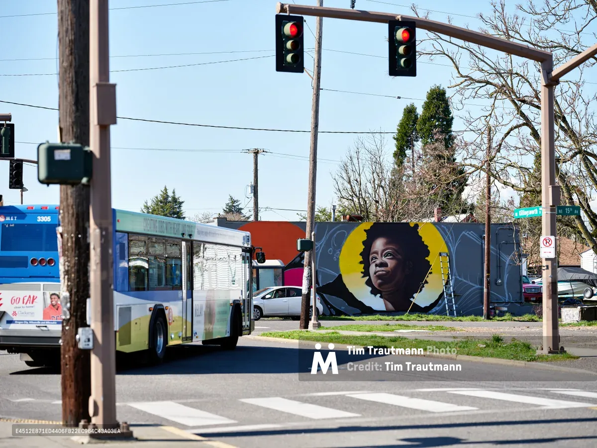 A vibrant street art portrait of an African American woman with natural hair dominates a wall along Northeast Killingsworth in Portland's historically Black Alberta neighborhood. The mural features bold golden circular elements framing the subject's contemplative expression against a blue-grey background. A TriMet bus pauses at the intersection under traffic lights, while bare winter trees and residential buildings complete this slice of Portland's evolving urban landscape.