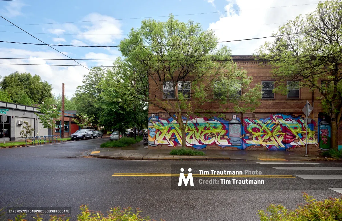 Vibrant graffiti-style murals cascade across the brick facade of a two-story building on Southeast Division Street, marking the former site of Portland's Oregon Theater. The kaleidoscopic artwork, featuring bold yellows, blues, and reds in dynamic letterforms, breathes new life into the Richmond neighborhood streetscape. Mature trees frame the scene under a partly cloudy spring sky, while power lines stretch overhead and modest commercial buildings anchor the quiet residential street.