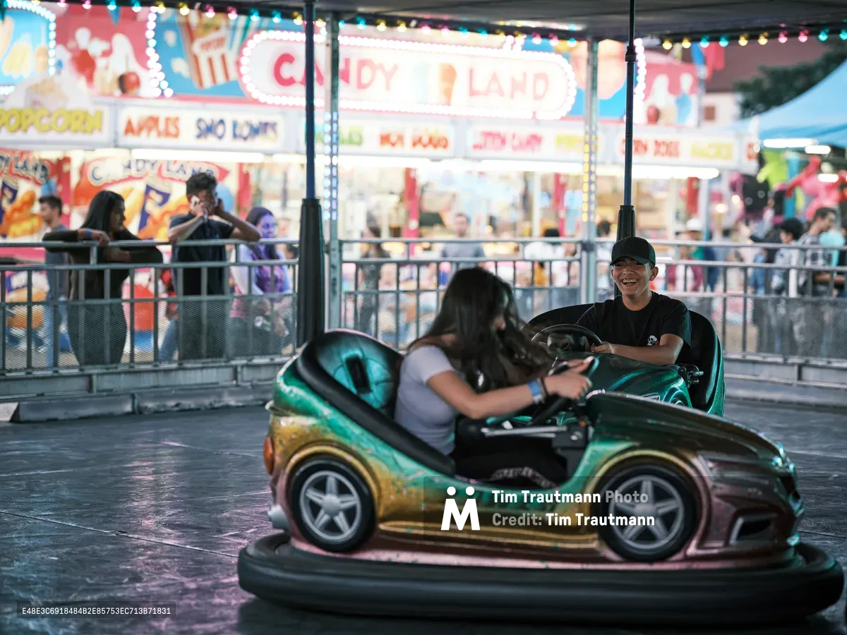 A young man grins with delight as he navigates a green and gold bumper car at the Clark County Fair in Vancouver, Washington. The iridescent vehicle catches the ambient carnival lighting while a passenger leans forward, both immersed in the classic fairground experience. Behind them, the bustling midway glows with colorful game booths and candy stands, their neon signs creating a vibrant backdrop of pure Americana. The scene captures the timeless magic of county fair entertainment under the gentle evening light.