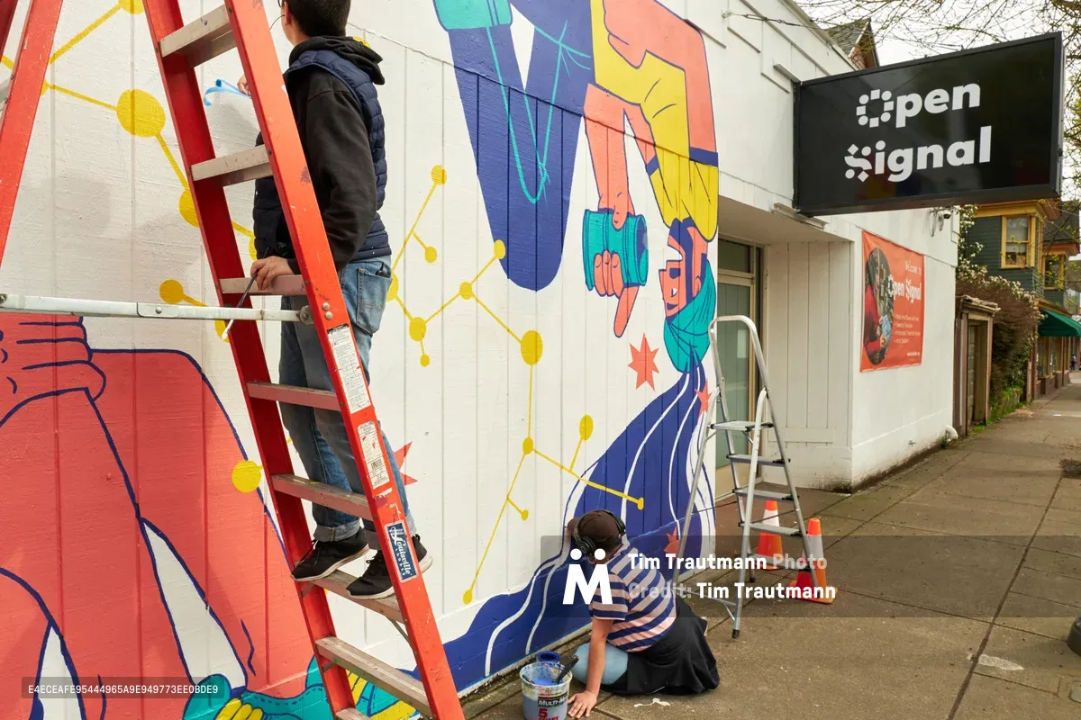 Two artists work in tandem on a vibrant public mural titled 'Let's Talk' outside Open Signal on Northeast Martin Luther King Jr. Boulevard in Portland's Eliot neighborhood. One artist perches on a red ladder applying paint to geometric patterns while another kneels below, adding details to flowing blue forms. The contemporary mural features bold oranges, blues, and yellows with interconnected nodes suggesting digital communication networks, transforming the white building facade into a canvas of community connection.