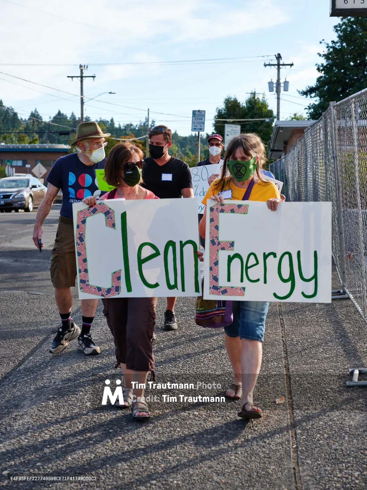 A diverse group of masked protesters marches along McLoughlin Promenade in Oregon City, carrying handmade banners spelling 'Clean Energy' in bold green lettering. The afternoon demonstration unfolds against a backdrop of power lines and suburban infrastructure, with participants ranging from an elderly bearded man in a fedora to young activists in casual summer attire. The scene captures the grassroots nature of environmental advocacy, as shadows stretch across weathered asphalt while demonstrators make their way toward Representative Kurt Schrader's office.