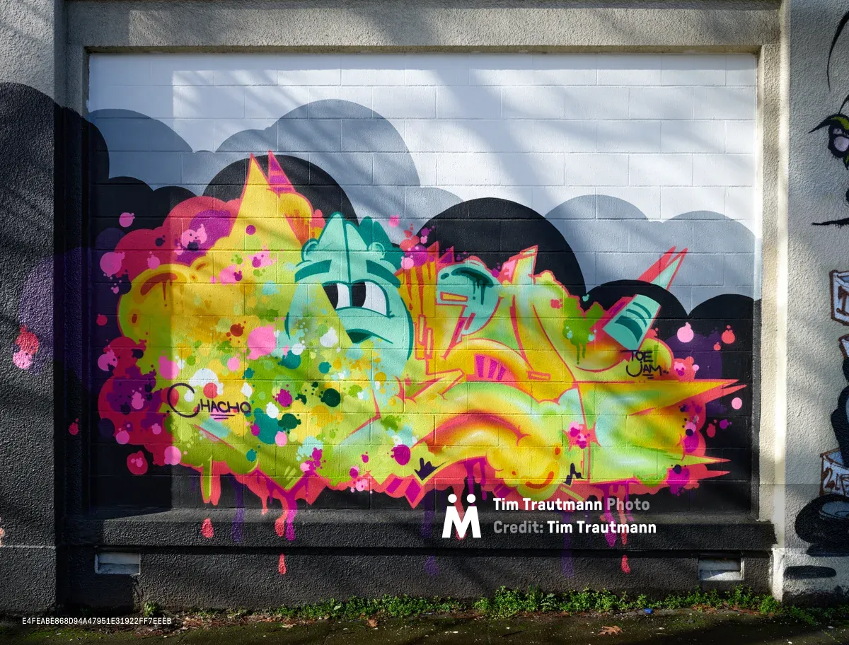 A kaleidoscopic mural explodes across white subway tiles on a former Salvation Army warehouse wall in Portland's Central Eastside district. The commissioned street art erupts in electric yellows, magenta pinks, and turquoise greens, with cartoon-like eyes peering through psychedelic clouds of color that seem to drip and flow beyond their boundaries. Natural daylight illuminates the vibrant composition, revealing the careful layering of spray paint techniques and the artist's signature embedded within the chromatic chaos.