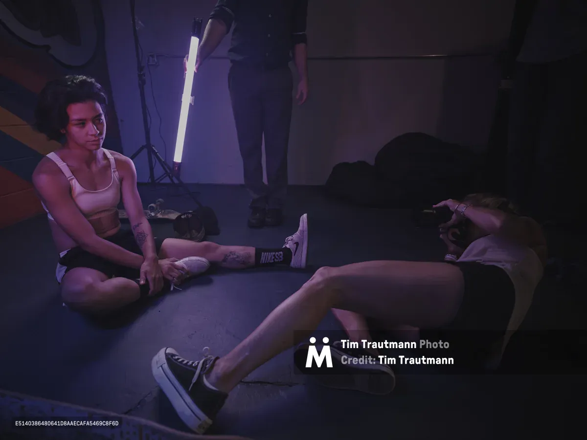 Professional skateboarder Christiana Means sits cross-legged on the concrete floor of Stronger Skatepark in Milwaukie, Oregon, her tattooed arms relaxed as she takes a break between sessions. The moody purple and magenta lighting from studio equipment casts dramatic shadows across the industrial space, while a photographer works nearby capturing the scene. The intimate moment captures the quiet intensity of skate culture, with Nike sneakers and scattered gear telling the story of dedicated practice.