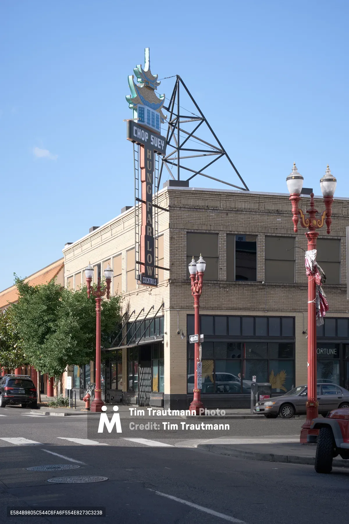 A vintage neon sign for "Chop Suey" rises above the historic brick buildings of Portland's Chinatown district, crowned with an ornate pagoda-style architectural element. The warm afternoon light bathes the multi-story commercial buildings while decorative red street lamps line the quiet intersection. Cars parked along the street and the clear blue sky create a peaceful urban scene that captures the enduring character of this culturally significant neighborhood.