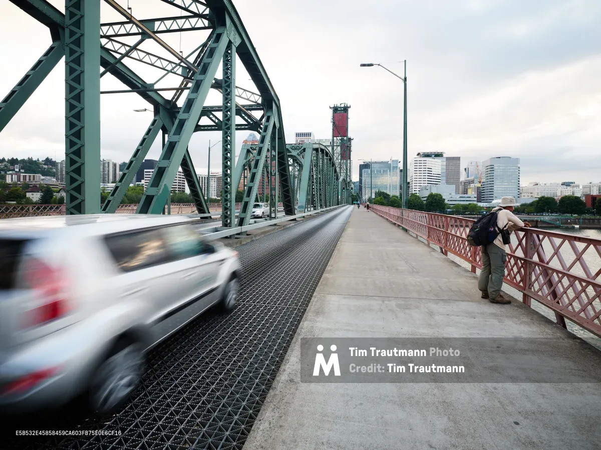 A solitary figure in a sun hat leans against the distinctive red pedestrian railing of Portland's iconic Hawthorne Bridge, gazing toward the city skyline as traffic streams past in a blur of motion. The weathered green steel truss structure frames the scene under overcast Pacific Northwest skies, while downtown Portland's modern towers rise beyond the Willamette River. The composition captures the contrast between human stillness and urban velocity, with the bridge's industrial architecture serving as a timeless connector between the city's east and west sides.