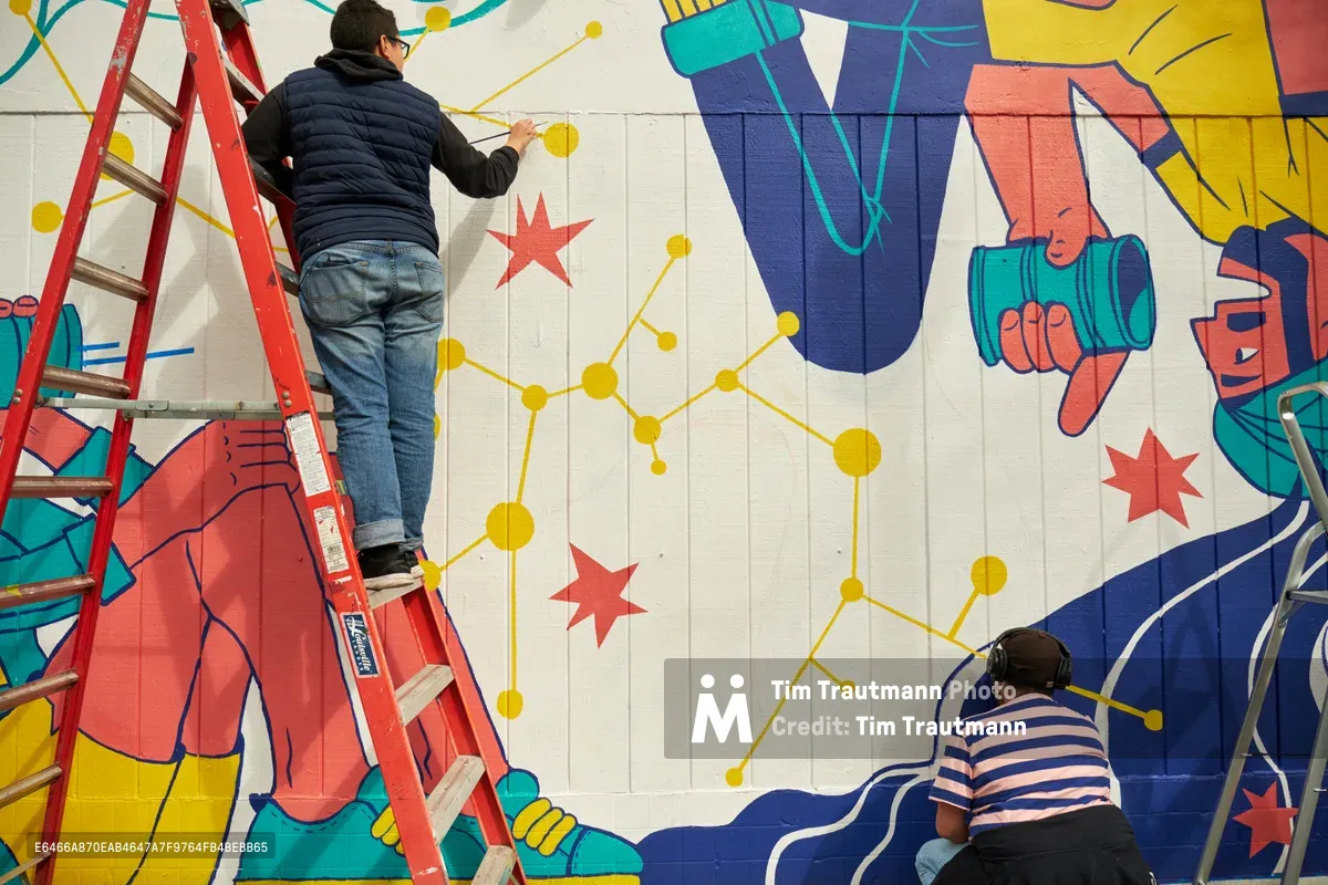 Two muralists work collaboratively on an expansive public art installation at Open Signal in Portland's Eliot neighborhood. One artist balances on a red ladder while adding delicate yellow constellation-like details to the vibrant composition, while another works at ground level on the lower portion of the wall. The mural features bold geometric forms in coral, turquoise, and navy blue, interwoven with red stars and connecting golden lines that create a sense of cosmic unity across the white concrete block surface.