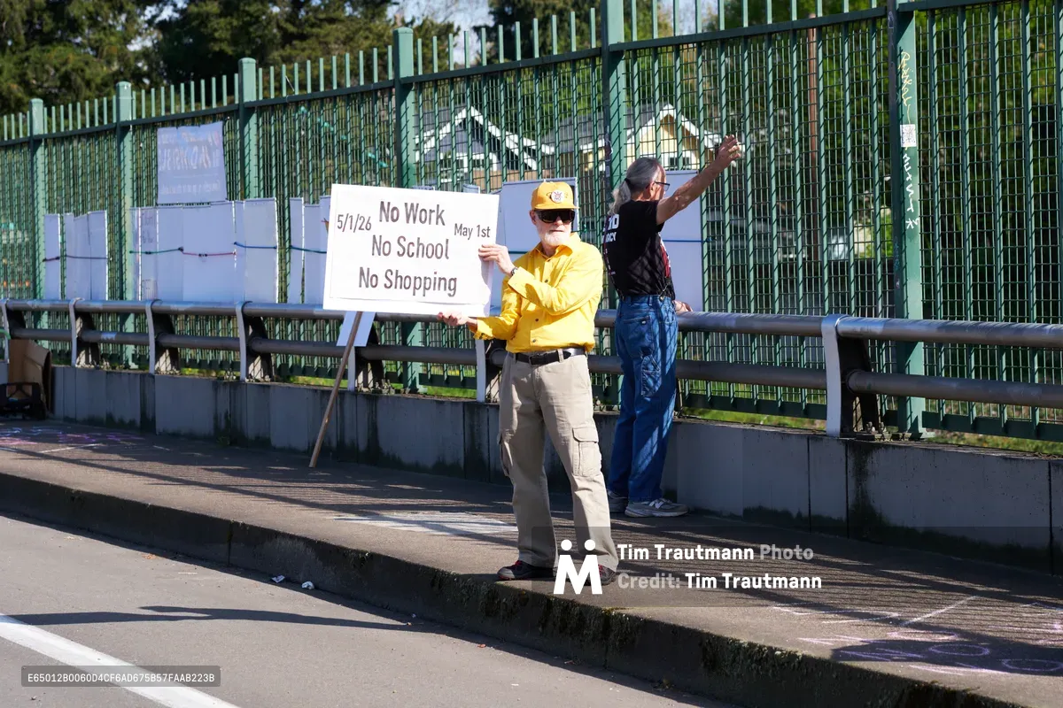 Two activists stand on a concrete pedestrian overpass above Interstate 5 in North Portland's Humboldt neighborhood, displaying a handwritten protest sign calling for a May 1st general strike. The man in a yellow shirt and baseball cap holds the white placard reading 'No Work No School No Shopping May 1st' while his companion in denim overalls raises his arm toward passing traffic below. Golden afternoon light casts long shadows across the bridge's weathered concrete surface, while green metal fencing and industrial structures frame this moment of grassroots political organizing. The urban landscape stretches behind them, punctuated by mature trees and the geometric patterns of highway infrastructure.