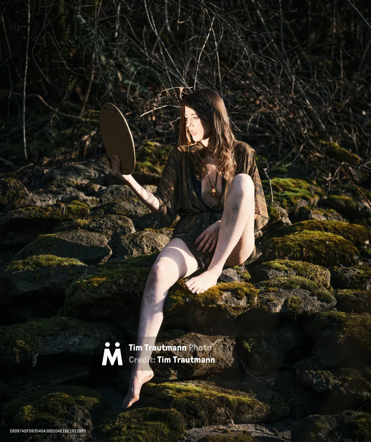 A young woman sits barefoot on moss-covered volcanic rocks along a riverbed on Oregon's Floodplain Trail, holding a circular mirror that catches the golden afternoon light. Her flowing brown hair and earth-toned dress harmonize with the ancient forest setting of bare winter branches and emerald moss. The warm sunlight creates dramatic shadows across the rocky terrain, while she gazes thoughtfully into the distance, creating an ethereal moment of self-reflection in nature.