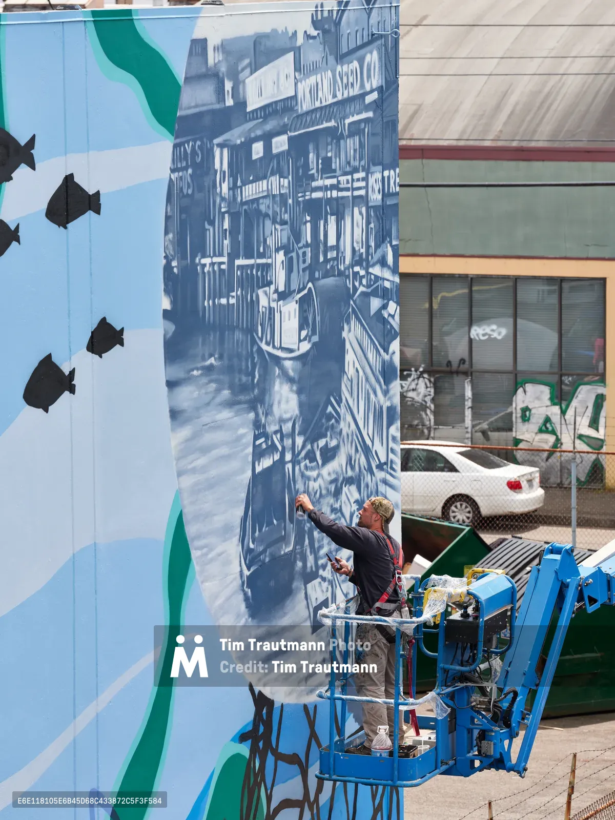 Jeremy Nichols and Devin Finley work from a blue hydraulic lift, adding intricate details to a large-scale monochromatic mural depicting historic streetscapes in Southeast Portland. The massive wall art transforms an urban corridor along Southeast 2nd Avenue in the Central Eastside district, where contemporary street art meets industrial architecture. Against a backdrop of chain-link fencing and parked vehicles, the artists methodically bring their vision to life under bright daylight, their collaborative effort representing the Portland Street Art Alliance's mission to enliven the city's built environment.