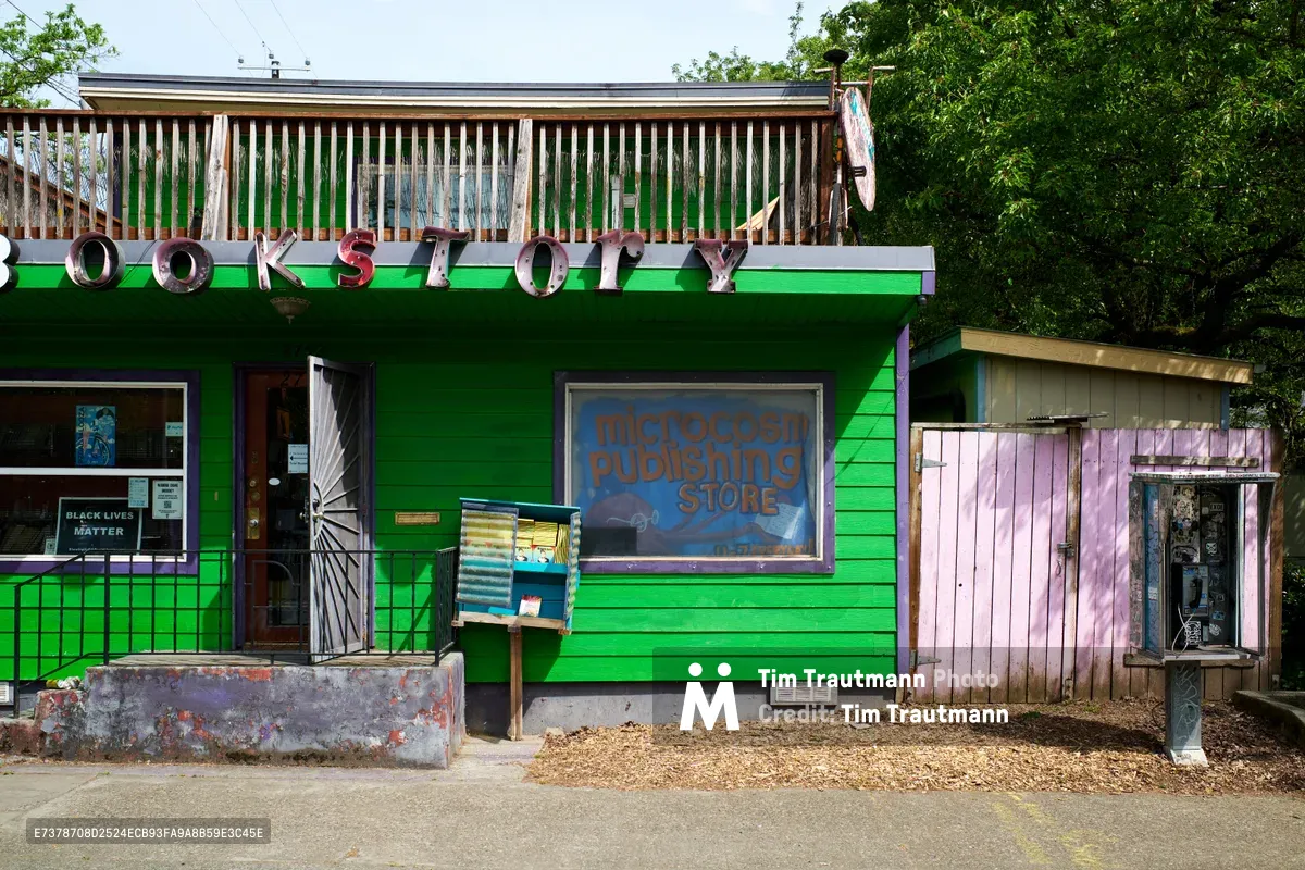 A striking emerald green wooden building houses Microcosm Publishing Store on North Williams Avenue in Portland's historically significant Eliot neighborhood. The rustic metal lettering spelling "BOOKSTORE" crowns the structure's upper balcony, while weathered wood siding and purple accents create an eclectic bohemian atmosphere. A public pay phone stands beside the entrance, embodying the grassroots community spirit of this Albina district establishment.