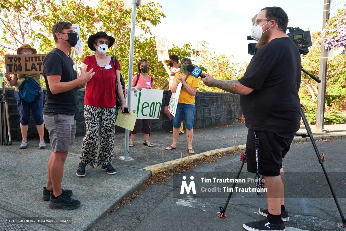 Michael Gaskill stands in animated conversation with a KOIN Channel 6 news crew on the sun-dappled sidewalk outside Representative Kurt Schrader's Oregon City office. Behind him, masked demonstrators hold handmade signs including "Too Little Too Late" and "Clean," their colorful clothing creating a vibrant backdrop against the autumn foliage. The golden afternoon light filters through leafy trees, casting warm shadows across the concrete as the interview unfolds during this climate change protest.