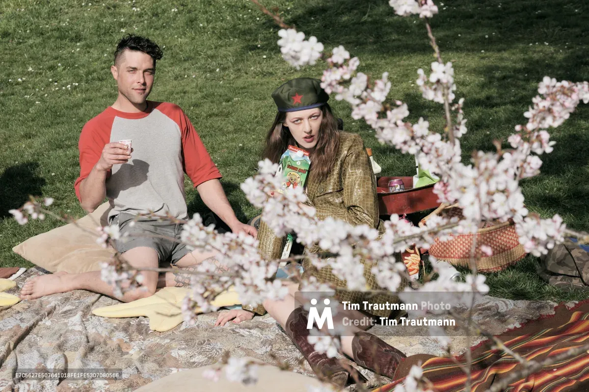 Two models pose for an editorial photography session beneath blooming cherry trees at Mount Tabor Park in Portland, Oregon. The woman, wearing a vintage military-style cap and golden jacket, sits alongside a man in a coral baseball tee as delicate pink and white blossoms frame the foreground. Soft afternoon light filters through the canopy, casting gentle shadows across their blanket spread with snacks and drinks on the verdant hillside.
