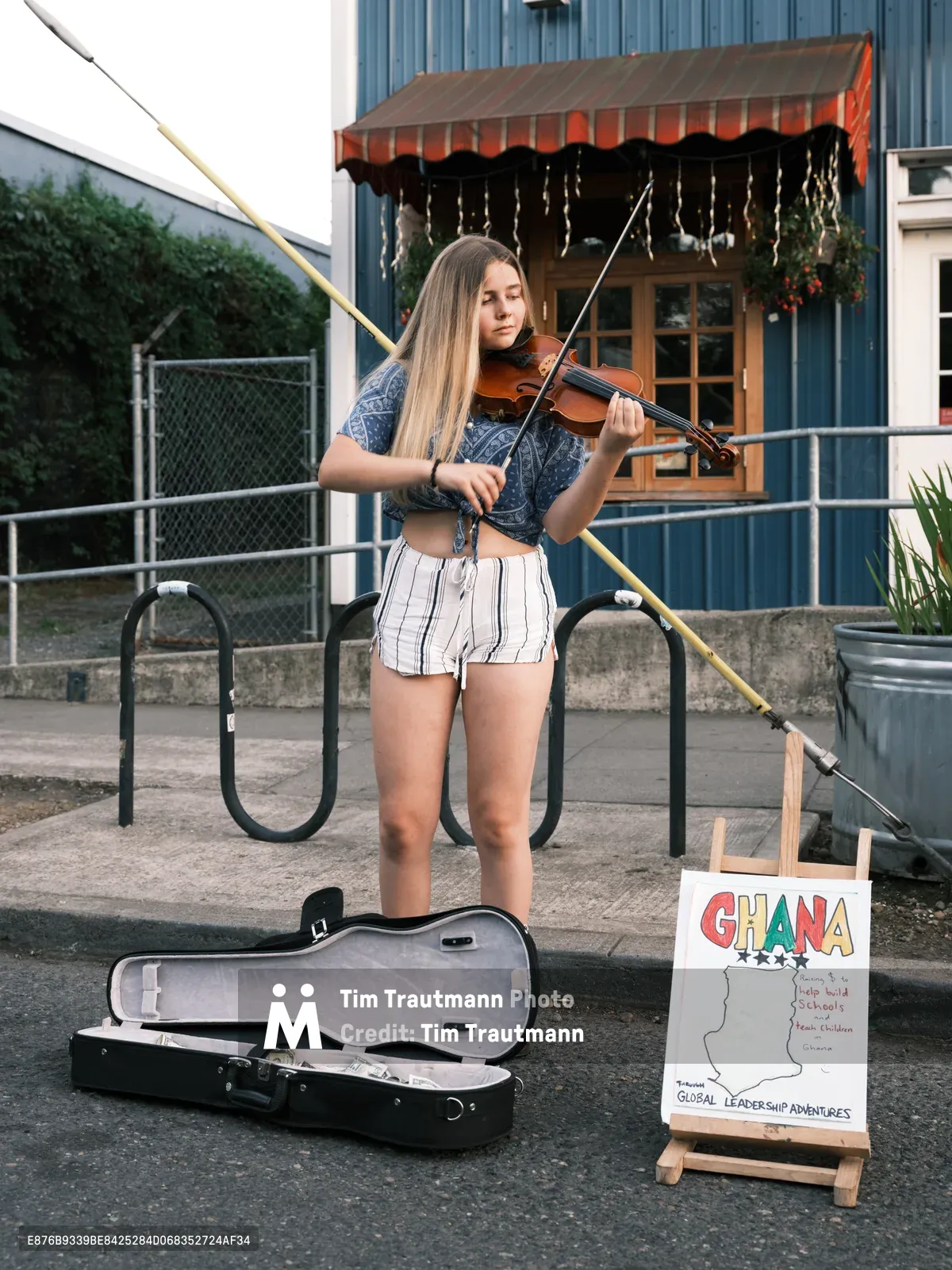 A young blonde woman performs violin on Northeast Alberta Street during Portland's Last Thursday street fair, her instrument case open for donations beside a colorful Ghana-themed sign. The late afternoon light catches the blue corrugated siding of the Waffle Window behind her, while she stands confidently in casual summer attire—denim top and striped shorts—bringing classical music to the bustling neighborhood gathering. Her setup includes a small easel displaying information about Global Leadership Adventures, suggesting a fundraising performance amid the community celebration.