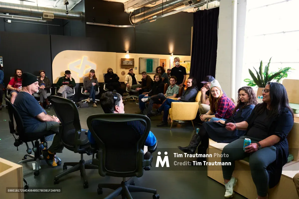 A diverse group of technology enthusiasts and developers congregate in an industrial-chic studio space in Portland's Ladd's Addition neighborhood for an ATProto meetup. The exposed ductwork and polished concrete floors frame an intimate circle of participants seated on modern furniture, with warm accent lighting creating a welcoming atmosphere against the dark feature wall. Black curtains and large windows define the CodeTV studio's professional yet relaxed environment.