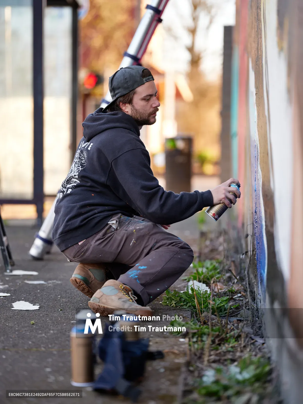 Travis Fields, known as campographic, crouches in focused concentration as he works on a mural depicting poet Amanda Gorman at the intersection of Northeast 15th Avenue and Killingsworth Street in Portland's Alberta district. The artist, wearing a dark hoodie and backwards cap, grips a spray paint can while surrounded by an array of colorful aerosol cans scattered on the asphalt. Golden afternoon light filters through the urban streetscape, casting warm tones across the scene as Fields transforms the concrete wall into a tribute to the celebrated inaugural poet.