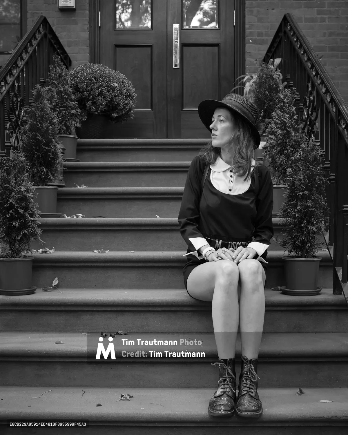 A young woman in vintage-inspired attire sits contemplatively on weathered stone steps, her gaze cast thoughtfully to the side beneath the shadow of a wide-brimmed hat. The monochromatic composition captures the intimate solitude of urban autumn, with ornate iron railings framing seasonal plantings that speak to Brooklyn's historic brownstone character. Fallen leaves scattered across the steps and her classic lace-up boots ground the scene in transitional seasonal beauty.