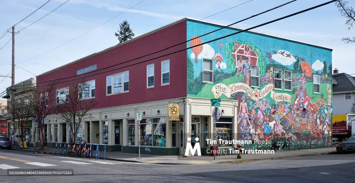 A vibrant corner building on Northeast Alberta Street showcases an elaborate community mural celebrating Portland's cycling culture. The two-story structure features burgundy upper siding contrasting with cream-colored ground-level storefronts, while the east-facing wall explodes with colorful artwork depicting hot air balloons, cyclists, and neighborhood scenes under the banner 'The Community Cycling Center.' Power lines crisscross the overcast sky above this quintessential Alberta District intersection, where bike share stations and urban cycling infrastructure reflect the neighborhood's commitment to sustainable transportation.