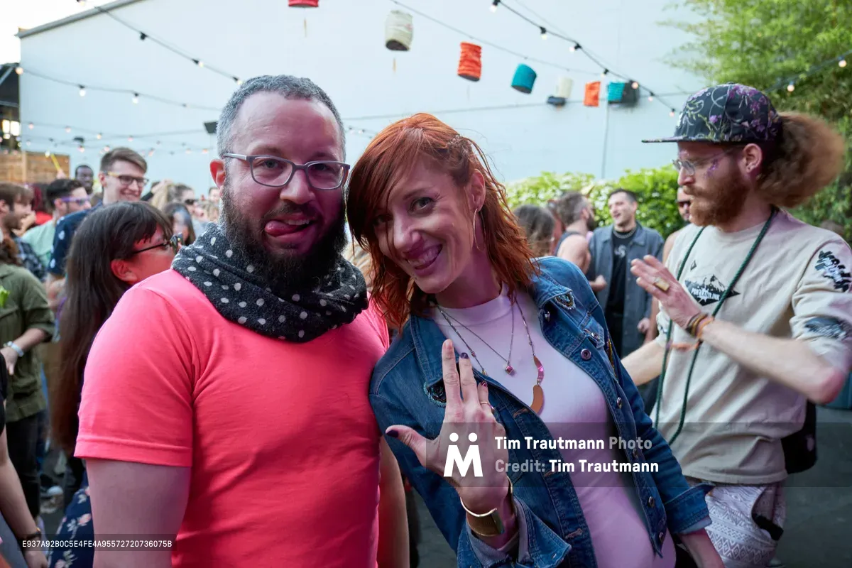 Two friends embrace the carefree spirit of Portland's summer day party scene under the dappled shade of White Owl Social Club's outdoor tent. The bearded man in coral pink and polka-dot bandana grins alongside a red-haired woman in denim who playfully flashes a peace sign, their joy infectious against the backdrop of fellow revelers. String lights overhead and colorful fabric bunting create an intimate festival atmosphere, while the crowd behind them pulses with the energy of Your Sunday Best's season opener.
