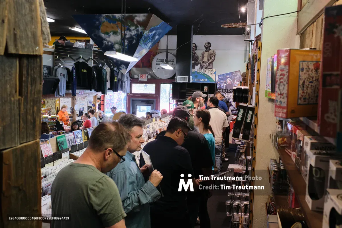 A bustling scene unfolds inside Music Millennium on Portland's East Burnside Street, where dozens of vinyl enthusiasts navigate the narrow, densely packed aisles of this legendary record store. The intimate space pulses with collectors hunched over crates, their faces illuminated by overhead fluorescent fixtures as they flip through countless album covers. Walls adorned with vintage posters and memorabilia create a nostalgic backdrop to this Saturday afternoon ritual of musical discovery.