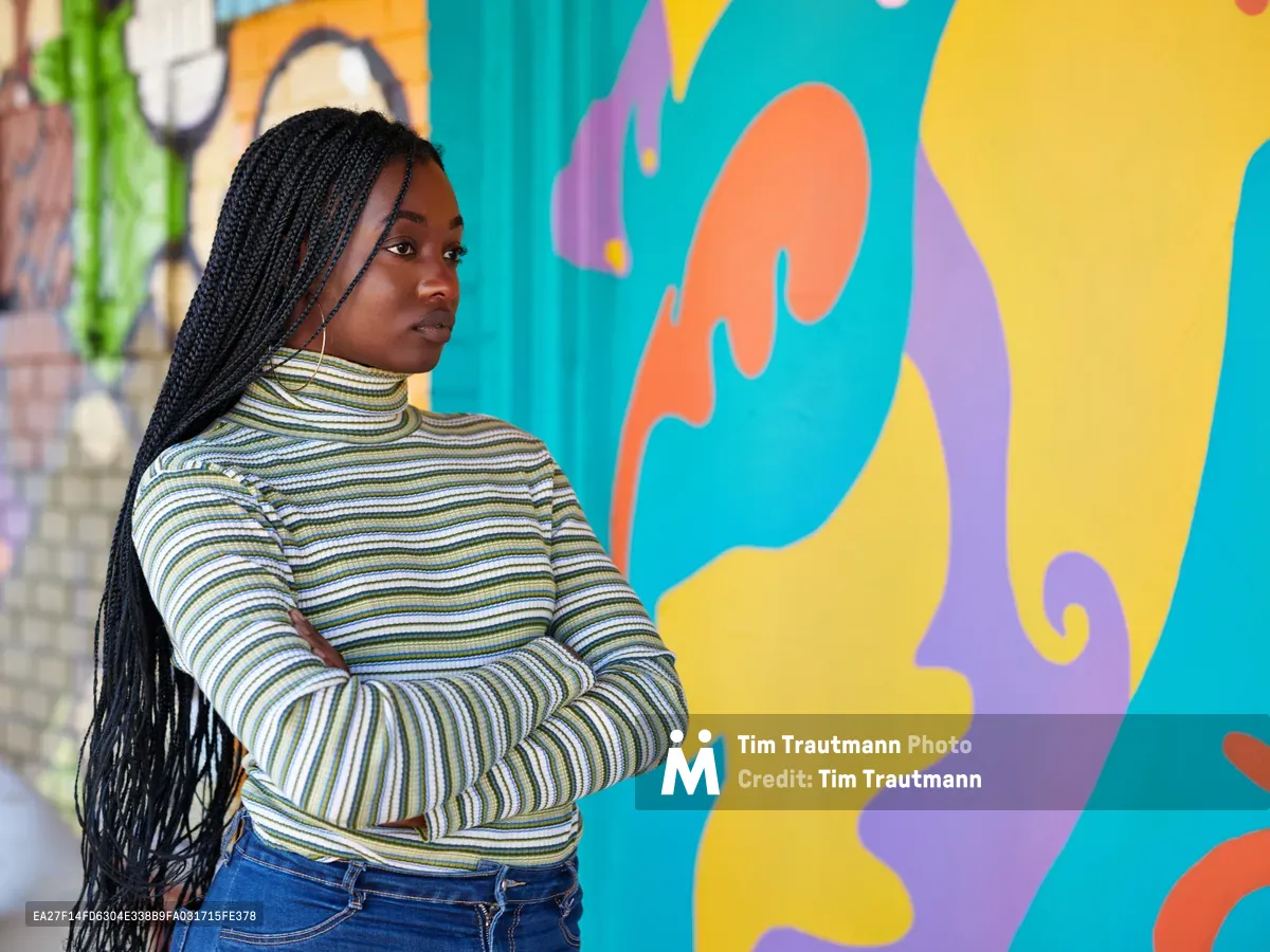 Naomi Likayi stands confidently before her completed mural in Portland's Alberta Arts District, her long braids cascading over a striped turtleneck as she poses with arms crossed. The dynamic street art behind her pulses with organic shapes in coral, turquoise, golden yellow, and violet, creating a bold backdrop that celebrates the neighborhood's creative renaissance. Soft daylight illuminates her contemplative expression, capturing a moment of artistic accomplishment against the kaleidoscopic wall that transforms the urban landscape.