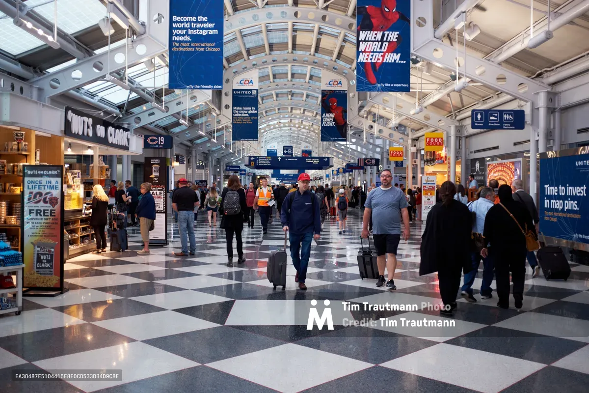 The iconic arched glass ceiling of Chicago O'Hare International Airport's terminal stretches overhead as travelers move through the bustling concourse with rolling luggage and backpacks. The distinctive black and white checkered floor creates geometric patterns beneath the steady flow of passengers, while suspended advertising banners and gate signage punctuate the soaring architectural space. Warm artificial lighting illuminates the commercial corridor, where retail storefronts line the walkway and directional signs guide the constant stream of travelers toward their destinations.