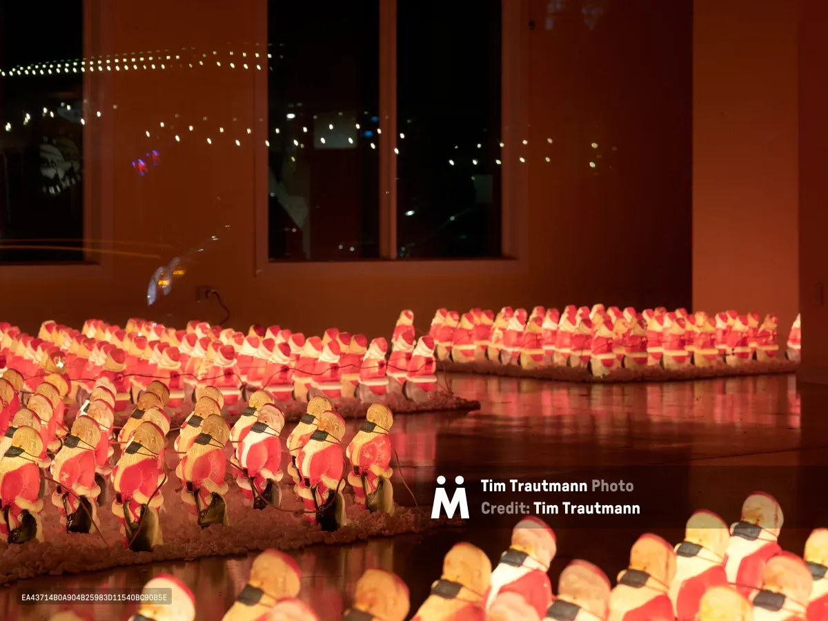 Rows of miniature Santa Claus figurines, each wearing black face masks, create a surreal holiday tableau behind the windows of a Sellwood-Moreland storefront in Portland, Oregon. The amber glow from interior lighting bathes the countless Santa clones in warm light, their red suits and white beards creating a sea of festive repetition. City lights twinkle through the glass, reflecting off the polished surface below the display, while the masked faces transform this traditional Christmas imagery into a poignant commentary on pandemic-era celebrations.