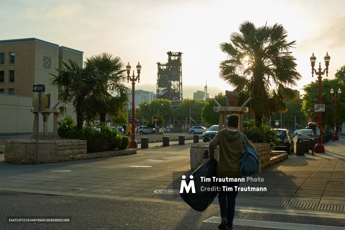 A solitary figure carries garbage bags across Northwest Everett Street in Portland's Chinatown-Japantown Historic District during the early morning hours. The soft dawn light filters through fan palms and illuminates ornate street lamps, while industrial structures and modern buildings create a layered urban backdrop. The scene captures the intersection of daily labor and historic neighborhood character, with the person silhouetted against the gentle, hazy sky that blankets this corner of Old Town Portland.