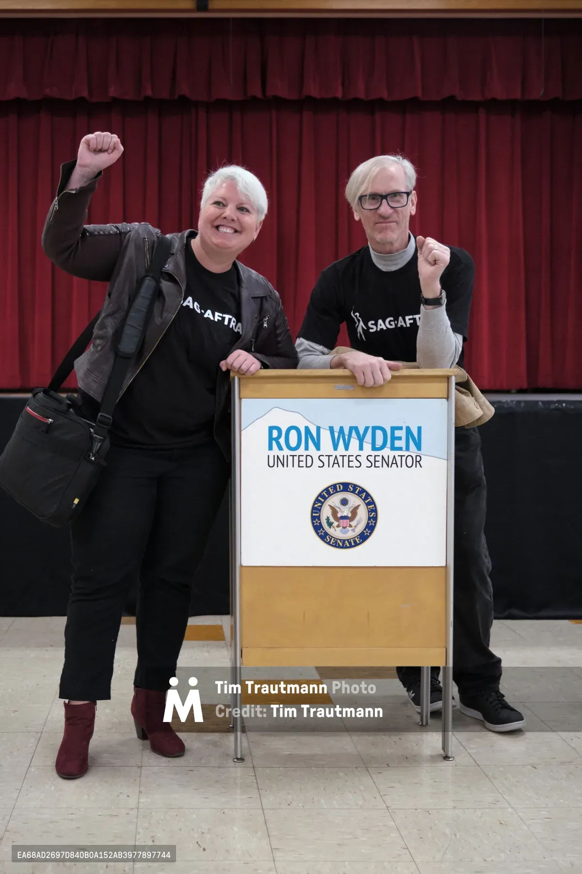 Two SAG-AFTRA union members stand triumphantly beside Senator Ron Wyden's podium at Robert Gray Middle School in Portland's Hillsdale neighborhood. The woman in a leather jacket raises her fist in solidarity while her companion gestures supportively, both wearing matching union t-shirts against the backdrop of burgundy curtains. The scene captures a moment of grassroots political engagement, with the official Senate podium creating a formal contrast to the intimate school auditorium setting.
