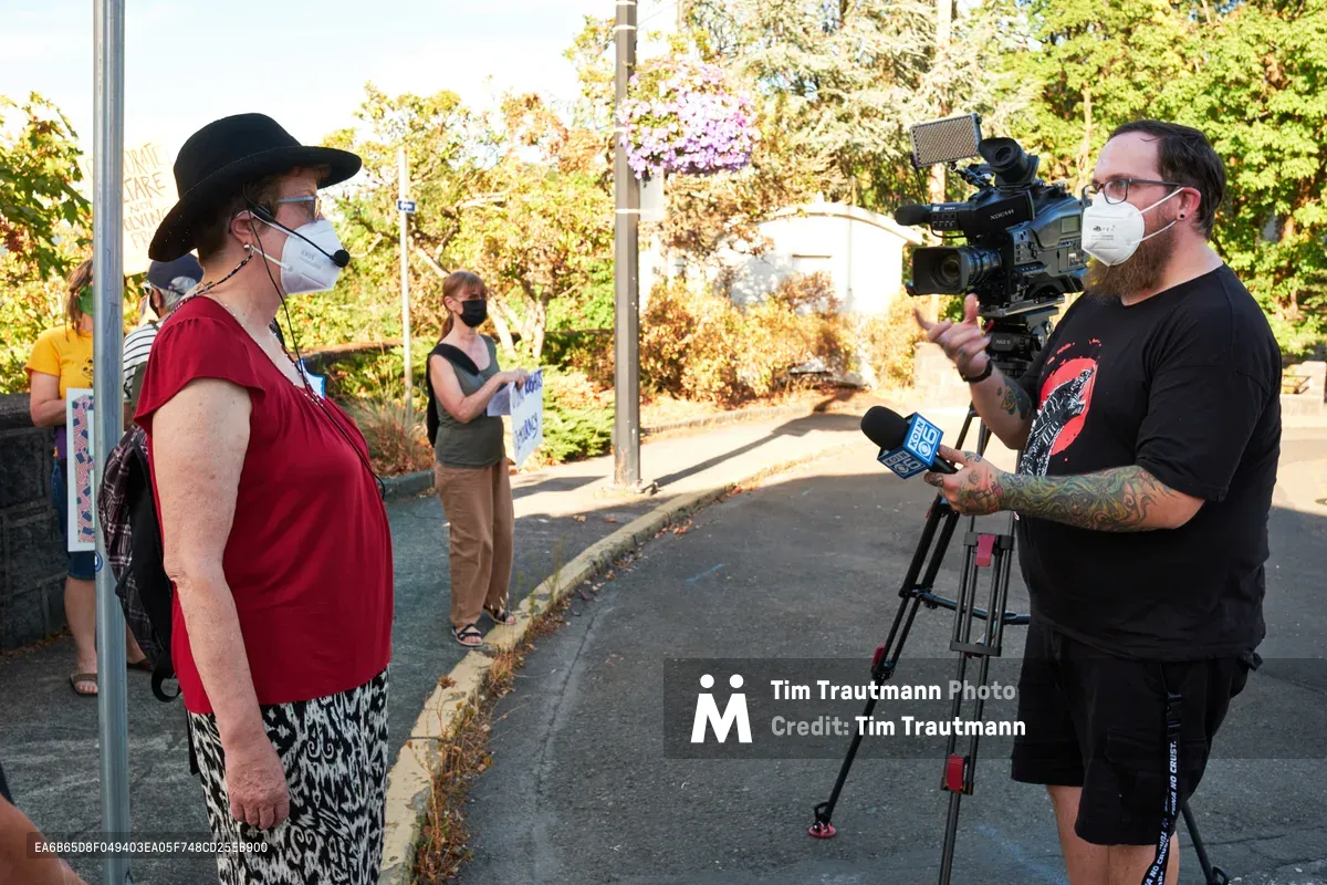 A masked climate activist in a red top and black hat speaks into a KOIN Channel 6 microphone during a rally outside Representative Kurt Schrader's Oregon City office. The bearded cameraman captures the interview against a backdrop of autumn foliage and suburban streets, while other masked demonstrators gather in the background. Golden afternoon light filters through the trees, creating a warm glow over this moment of grassroots political engagement.