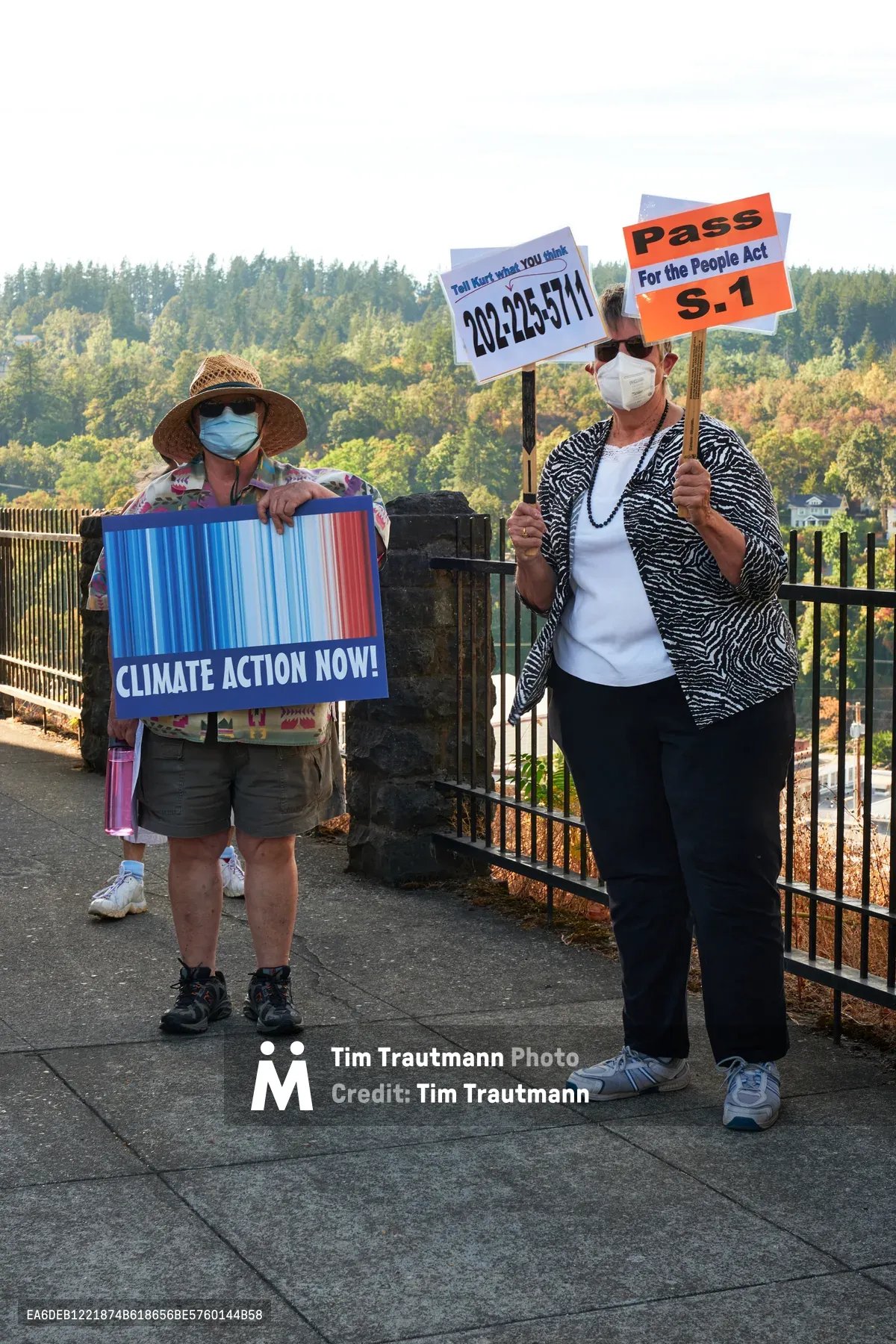 Two masked protesters stand on concrete pavement outside Representative Kurt Schrader's Oregon City office, holding colorful signs advocating for climate action and legislative reform. The woman on the left displays a vibrant blue and red striped banner reading "CLIMATE ACTION NOW!" while wearing a woven sun hat and casual hiking attire. Her companion holds dual signs referencing phone numbers and "For the People Act S.1" legislation. Behind them, autumn foliage creates a warm backdrop along the McLoughlin Promenade, with golden and rust-colored trees framing the civic demonstration in soft, overcast light.