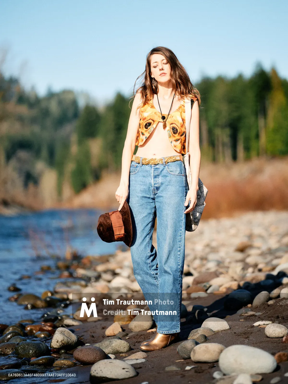 A young woman with flowing brown hair stands contemplatively along the rocky river bed of the Columbia River in Multnomah County, Oregon. She wears a distinctive animal-print crop top paired with high-waisted denim and carries a brown leather bag, her eyes gently closed as golden hour light illuminates her face. The river's gentle waters lap against smooth river stones while evergreen forests create a soft backdrop under the clear Pacific Northwest sky. Her bohemian styling and peaceful demeanor capture a moment of serene connection with Oregon's natural landscape.