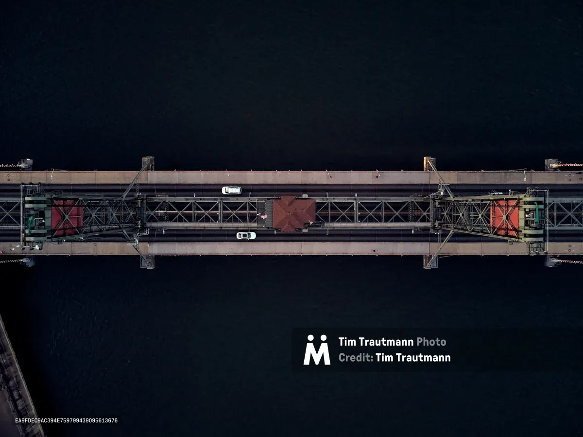 A precise aerial perspective captures Portland's historic Hawthorne Bridge spanning the dark Willamette River at dusk. The steel truss structure stretches across the frame in perfect symmetry, its weathered concrete deck illuminated by scattered streetlights while two vehicles traverse the span. The inky waters below create a stark void that emphasizes the bridge's engineered geometry and vital connection between Portland's eastside neighborhoods and downtown core.