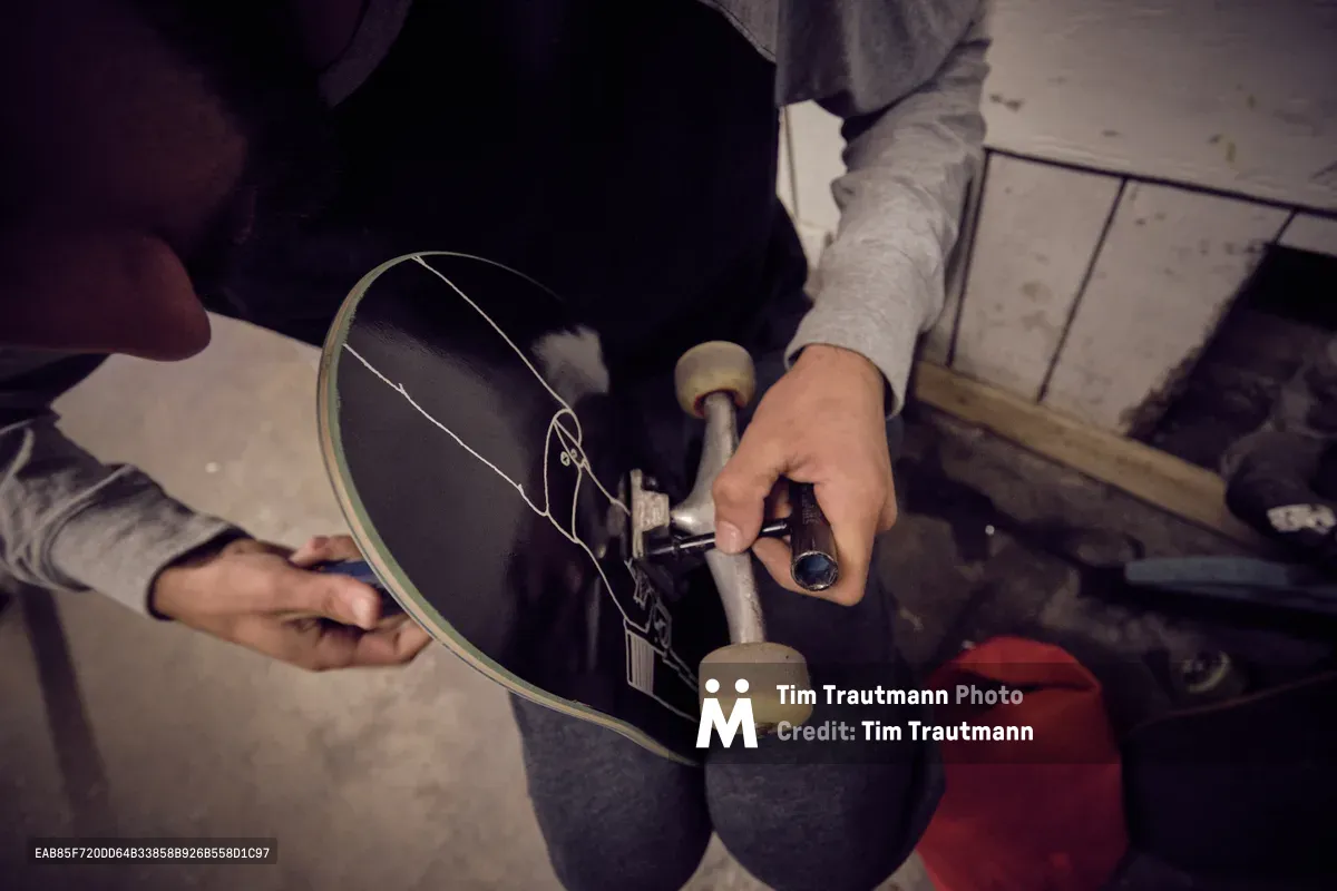 In the intimate confines of Commonwealth Skateboarding on Southeast Portland's 20th Avenue, hands work methodically over a black skateboard deck, adjusting trucks with practiced precision. The scene unfolds in warm, muted light that casts gentle shadows across weathered concrete floors, while red and gray hoodies frame the focused maintenance ritual. This quiet moment captures the behind-the-scenes dedication that keeps Portland's skate culture rolling.