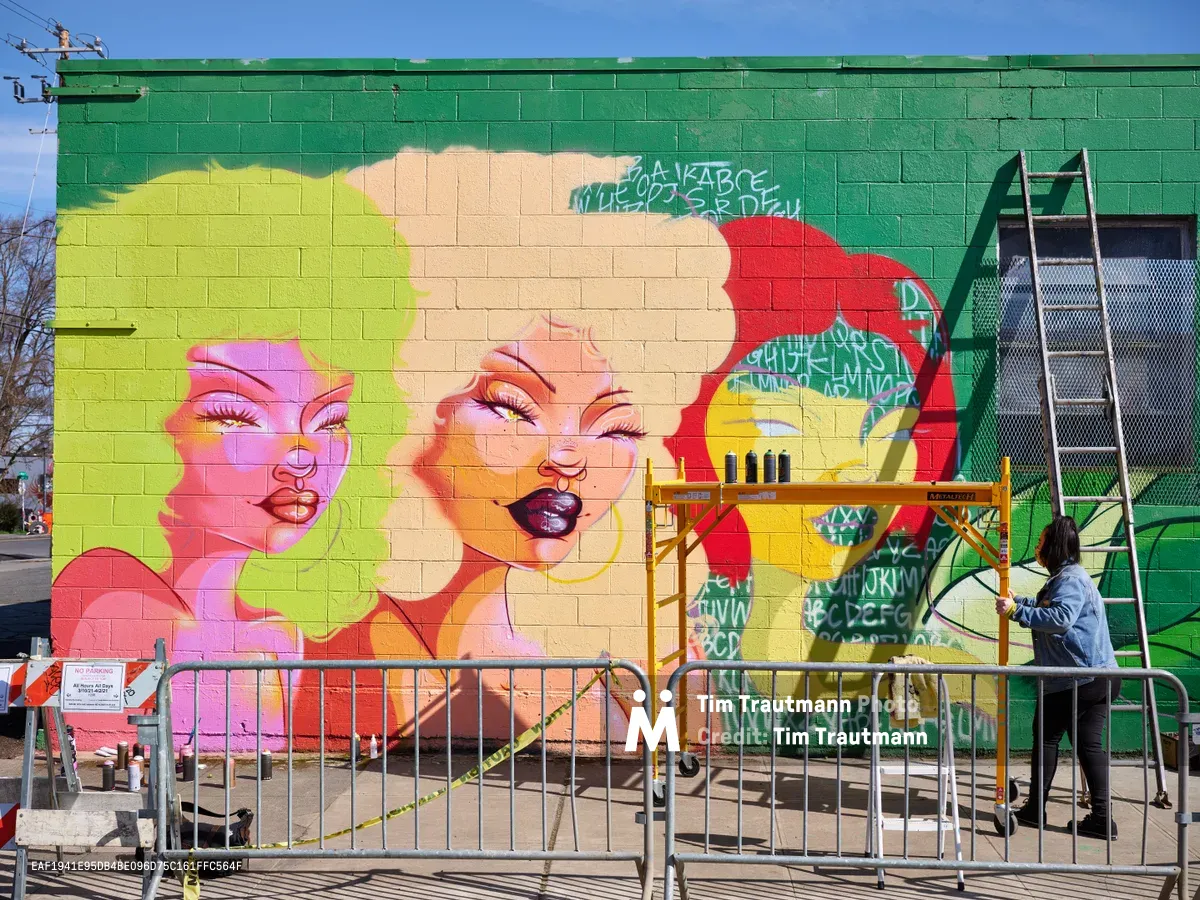 Multidisciplinary artist Vilatose works on 'Jardín Cubana,' a striking mural featuring three stylized female figures with vivid pink, yellow, and red hair against an emerald green cinderblock wall. The scene captures the creative energy of Portland's Alberta Arts District, with construction barriers and scaffolding framing the artist at work under clear blue skies. The mural showcases bold feminine portraits rendered in contemporary street art style, transforming an urban wall into a canvas of color and expression.