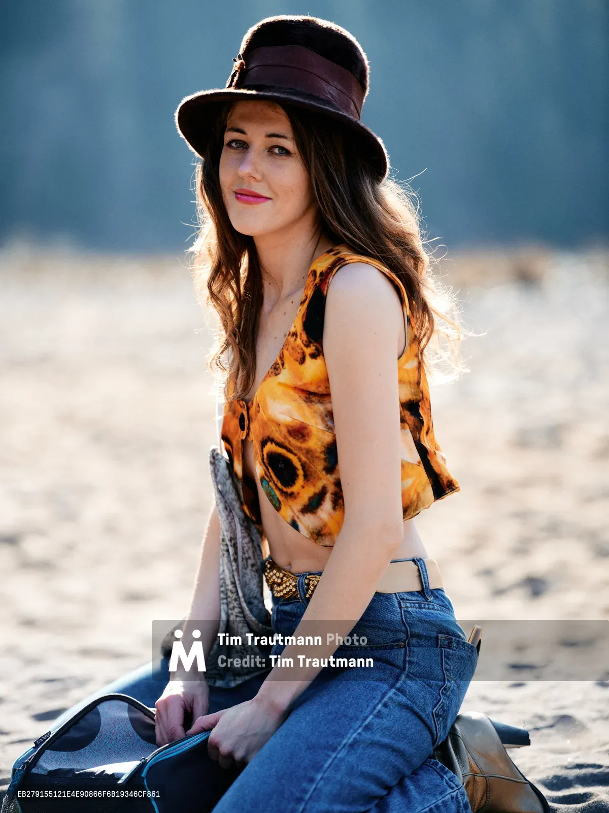 A young woman with flowing brunette hair sits gracefully on weathered driftwood along Oregon's Floodplain Trail, her vintage-inspired ensemble catching the warm afternoon light. She wears a distinctive orange floral crop top paired with high-waisted denim and a classic wide-brimmed hat with purple ribbon detail. The golden backlighting creates a luminous halo effect around her silhouette against the soft, neutral tones of the riverbed. Her confident pose and serene expression embody the carefree spirit of Pacific Northwest outdoor culture.