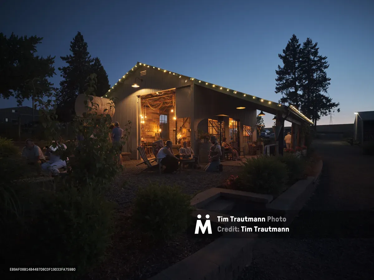 The exterior of the barn at Abbey Road Farm in Carlton, Oregon, photographed at dusk. The corrugated metal barn is strung with warm globe lights along its roofline, glowing invitingly against a deep blue twilight sky. Groups of guests relax on Adirondack chairs and benches on the gravel patio, socializing in the warm light spilling from the open barn interior. Tall evergreen trees flank the building, and lush garden plantings border a stone pathway in the foreground.