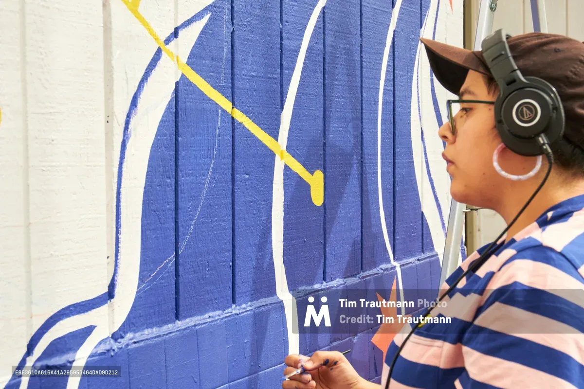 Maria Rodriguez, known as Sparkykneecap, works intently on a vibrant blue and yellow mural at Open Signal in Portland's Eliot neighborhood. The Mexican-American artist wears black headphones and a striped shirt while painting geometric shapes on the weathered wooden wall. Warm afternoon light illuminates the creative process as Rodriguez adds precise brushstrokes to the commissioned piece titled 'Let's Talk,' exploring themes of identity and culture through bold color and form.