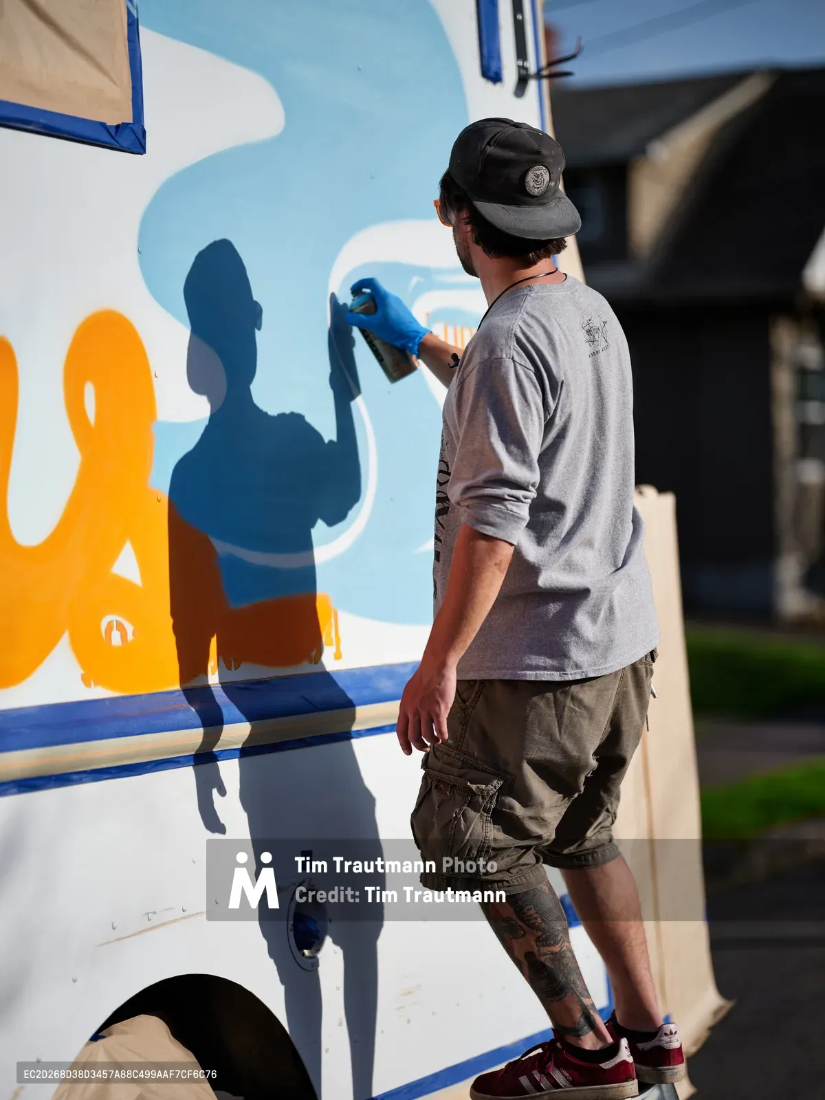 A muralist in a black baseball cap and gray long-sleeve shirt carefully applies vibrant blue paint to a food truck's exterior on Northeast 9th Avenue in Portland's King neighborhood. His tattooed leg and casual cargo shorts speak to the artistic street culture as he works under bright daylight, transforming the white vehicle with bold orange and blue graphics. The urban setting provides an authentic backdrop for this moment of creative commerce.